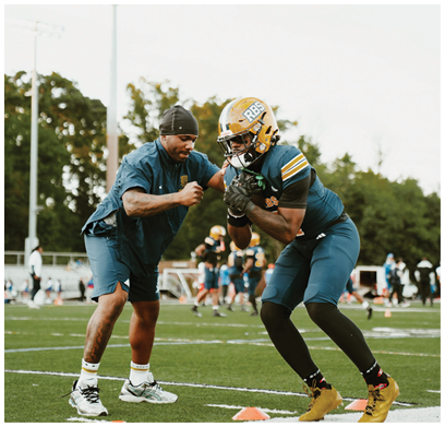 An adult wearing sports gear and a helmet is practicing football drills with a coach on a grassy field marked with orange cones. Description generated by AI