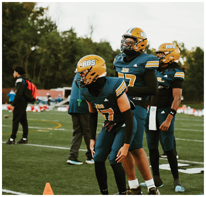 Three people wearing yellow helmets and dark sports uniforms stand on a field with trees in the background. Description generated by AI