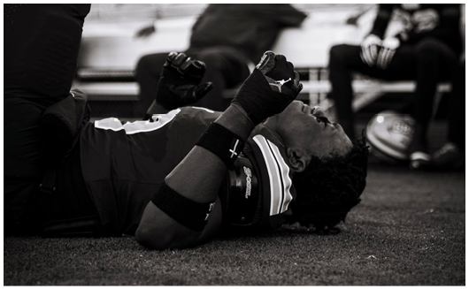 A person wearing sports gear lies on the ground with clenched fists raised, appearing to express strong emotion, while two others sit in the background near a soccer ball. Description generated by AI