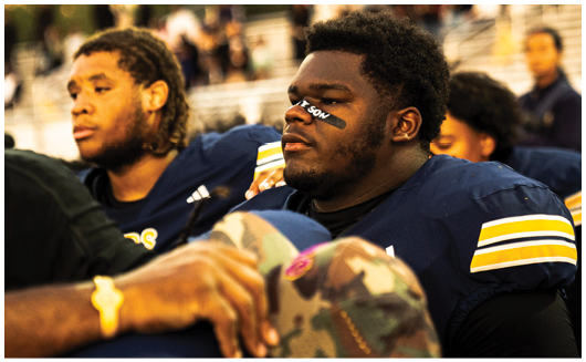 People in football uniforms sit closely together on a bench, with one person wearing eye black and looking focused. Description generated by AI