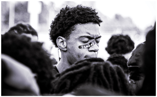 A person with short curly hair has a sticker on their face with the word “ISOLATE" while surrounded by a crowd of people in a black and white photo. Description generated by AI