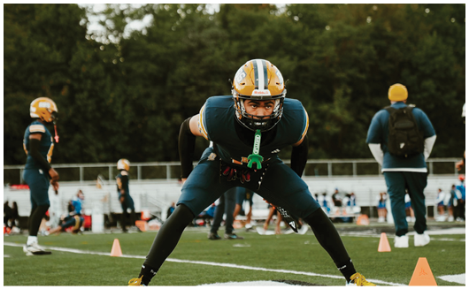 An adult in football gear is crouched in a ready stance on a field with other players and a coach in the background. Description generated by AI