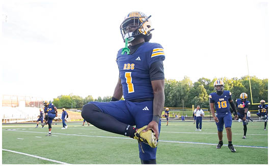 A person wearing a football helmet and uniform stands outdoors under a bright, partly cloudy sky. Description generated by AI