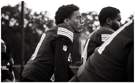 Three people in football uniforms are standing closely together outdoors, with one person in the foreground looking intently ahead. Description generated by AI