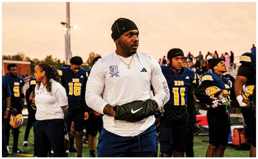 A group of people wearing sports attire stand on a field with some holding helmets and others looking focused, suggesting a team preparing for a game or practice. Description generated by AI