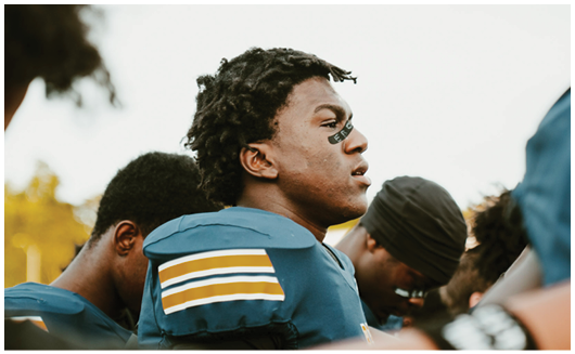 A person wearing a blue sports uniform with shoulder pads and eye black is standing among others in similar attire, appearing focused. Description generated by AI