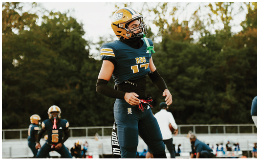 A person wearing a yellow helmet and sports gear is holding a football on a field with trees in the background. Description generated by AI