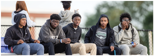 Five people wearing casual clothing sit on bleachers outdoors while two others stand in the background. Description generated by AI