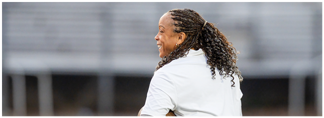 A person with curly hair tied back is smiling and looking to the side while wearing a white shirt in an outdoor setting. Description generated by AI