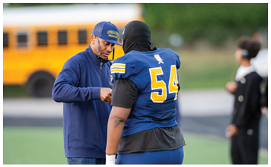 An adult wearing a blue jacket and cap is interacting with a person in a blue and yellow sports uniform with a blurred yellow school bus in the background. Description generated by AI