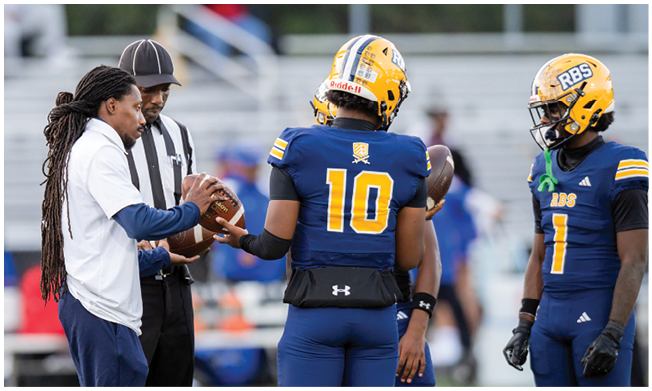 Three people wearing blue athletic pants stand on a sports field with one person’s hand resting on their thigh. Description generated by AI