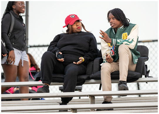 Two people sit on bleachers engaged in conversation, one wearing a red cap and black outfit and the other wearing a green and white jacket with beige pants. Description generated by AI