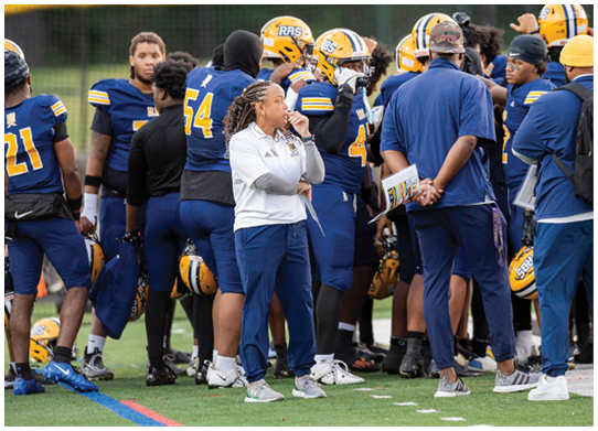 A group of people in sports uniforms and coaching attire are gathered on a field, appearing to discuss or strategize during a game or practice. Description generated by AI