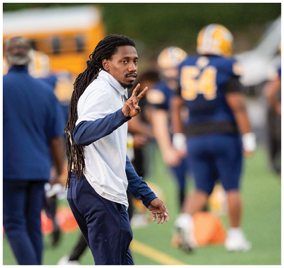 A person with long dreadlocks wearing a white jacket and navy sleeves is holding up three fingers while standing on a sports field with blurred players in blue and yellow uniforms in the background. Description generated by AI