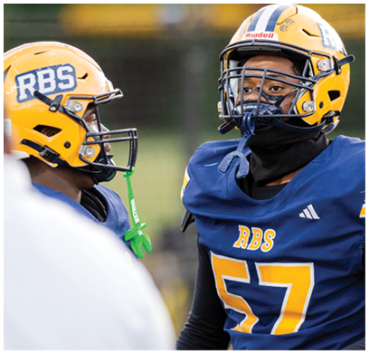 A person wearing a blue sports jersey with the number 57 and the letters RBS is standing next to another person in a white shirt. Description generated by AI