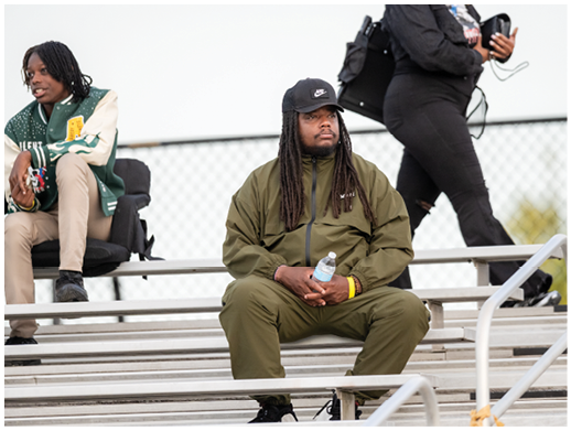 A person wearing a green tracksuit and black cap is sitting on metal bleachers holding a water bottle while another person walks by in the background. Description generated by AI