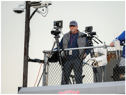 A person wearing a cap and jacket stands behind a chain-link fence surrounded by multiple cameras and electronic equipment. Description generated by AI