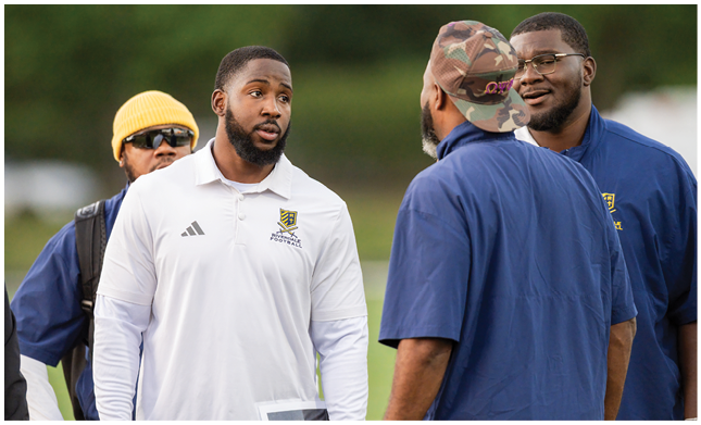 Four adults are engaged in conversation outdoors, with two wearing blue shirts and one in a white sports polo, while another wears a yellow beanie and sunglasses. Description generated by AI