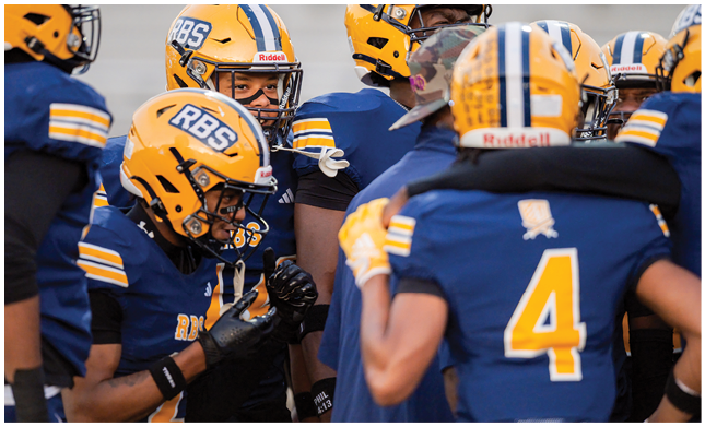 A group of people wearing yellow helmets and blue football uniforms are gathered closely, appearing to be in a team huddle or discussion. Description generated by AI