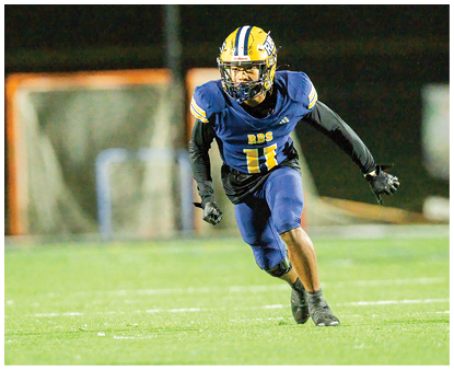 Two people in football gear are positioned on a field at night with blurred figures in the background. Description generated by AI