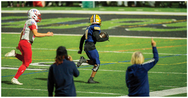 A person in a blue and yellow football uniform is running with a football while another person in a white and red uniform chases them on a sports field with two people watching from the sidelines. Description generated by AI
