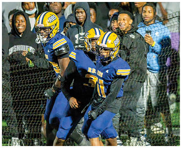 Two football players in blue and yellow uniforms are on the field near a net with a crowd of people watching behind them. Description generated by AI