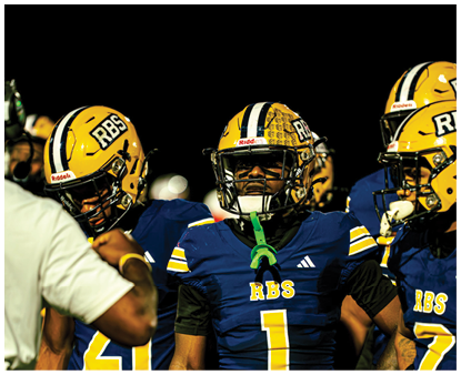 Two people in football gear are positioned on a field at night with blurred figures in the background. Description generated by AI