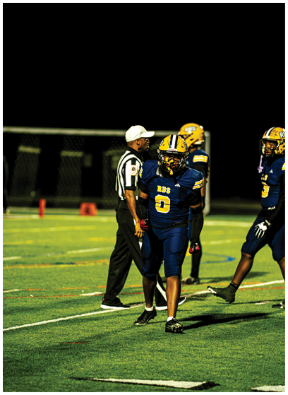 A person wearing a blue and yellow football uniform sits on a bench on a sports field with a helmet placed nearby. Description generated by AI