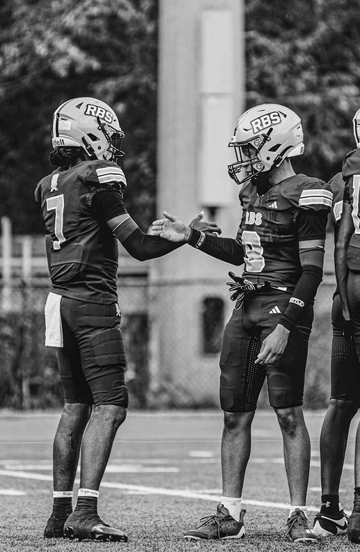 Two people wearing football uniforms and helmets are standing on a field facing each other and appear to be about to shake hands. Description generated by AI
