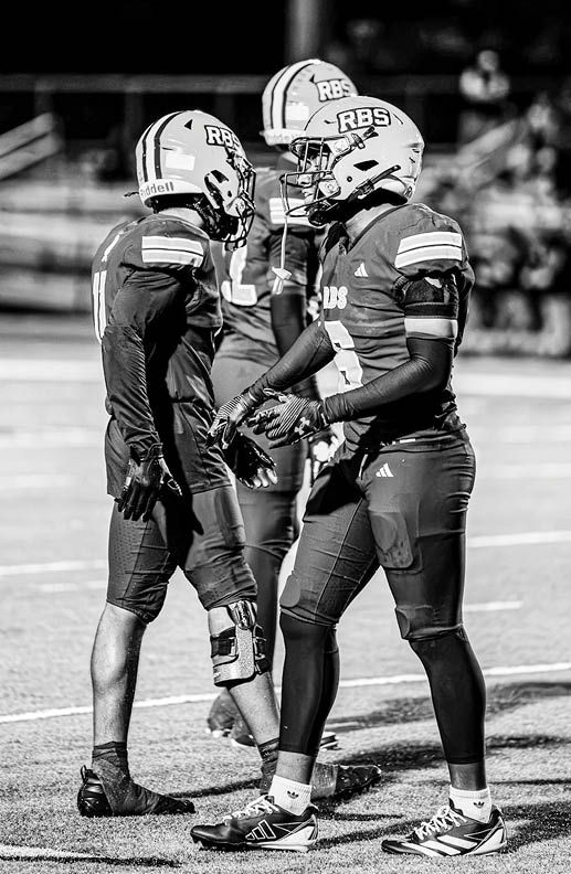 Two people in football uniforms and helmets are standing on a field facing each other, with one person wearing a knee brace. Description generated by AI