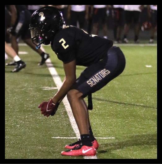 A person wearing a black football uniform with “VISTA" on the jersey is sitting on a grassy field with one leg bent and the other extended, next to a black football helmet. Description generated by AI