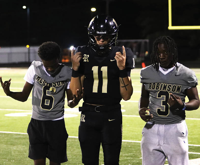 Four people wearing black football uniforms with “VISTA" on the front and face paint are standing together in a stadium with a crowd in the background. Description generated by AI