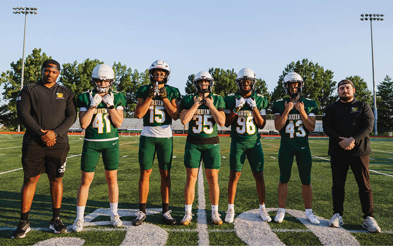 Six people wearing football uniforms and helmets stand on a football field with two others in casual sportswear standing on either side under a clear sky. Description generated by AI
