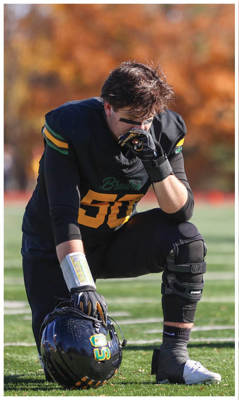 A person in a black football uniform with yellow and green accents kneels on a field holding a helmet and appears to be wiping their face. Description generated by AI