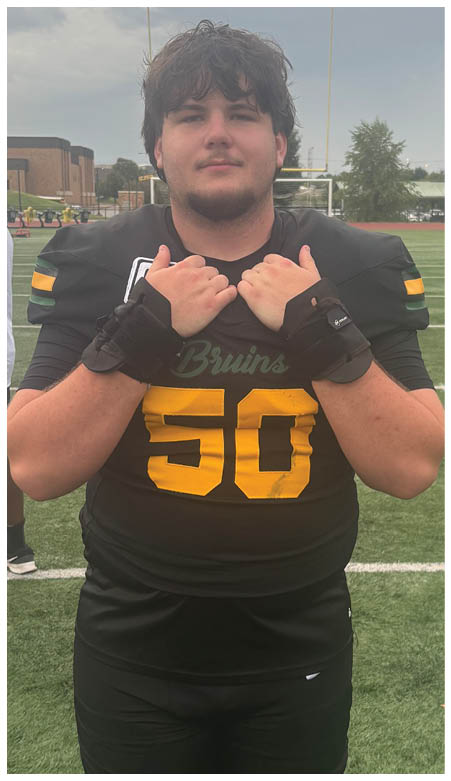 A person wearing a black football jersey with the number 50 and the word Bruins stands on a football field holding the collar of the jersey with both hands. Description generated by AI