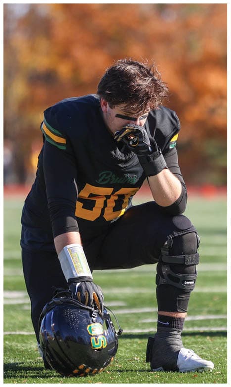 A person in a black football uniform with yellow and green accents kneels on a field holding a helmet and appears to be wiping their face. Description generated by AI