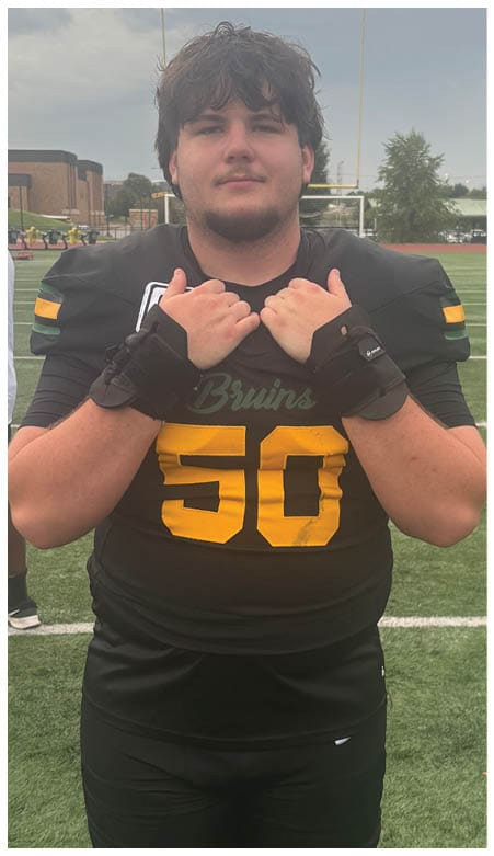 A person wearing a black football jersey with the number 50 and the word Bruins stands on a football field holding the collar of the jersey with both hands. Description generated by AI