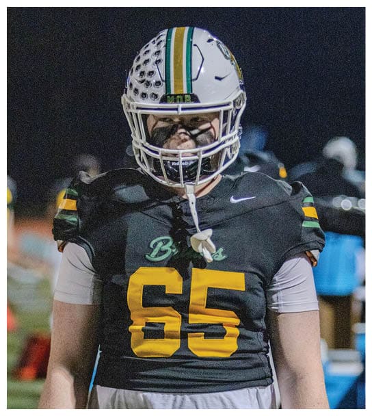 A person wearing a football uniform with the number 65 and a helmet stands on a field at night. Description generated by AI