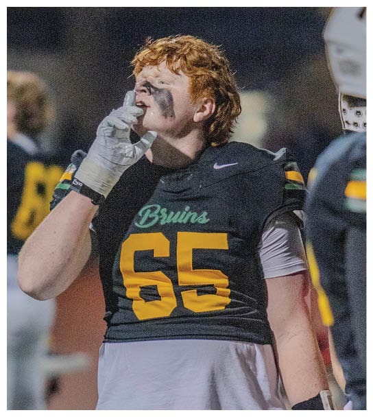 A person with curly hair wearing a football uniform with “Bruins" on the chest is drinking from a water bottle during a nighttime game. Description generated by AI