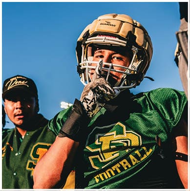 A person wearing a football helmet and green jersey gestures with a gloved hand while two other people stand nearby against a clear blue sky. Description generated by AI