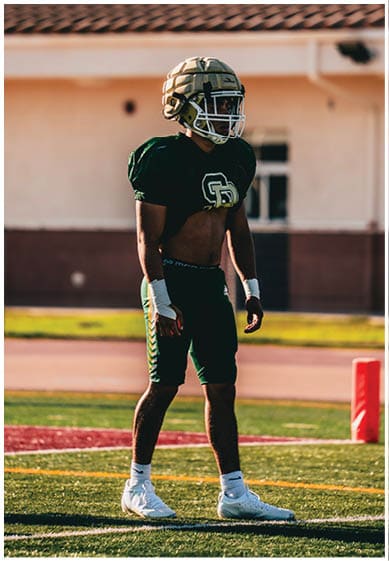 A person wearing a football helmet and uniform stands on a field near a red end zone marker with a building in the background. Description generated by AI