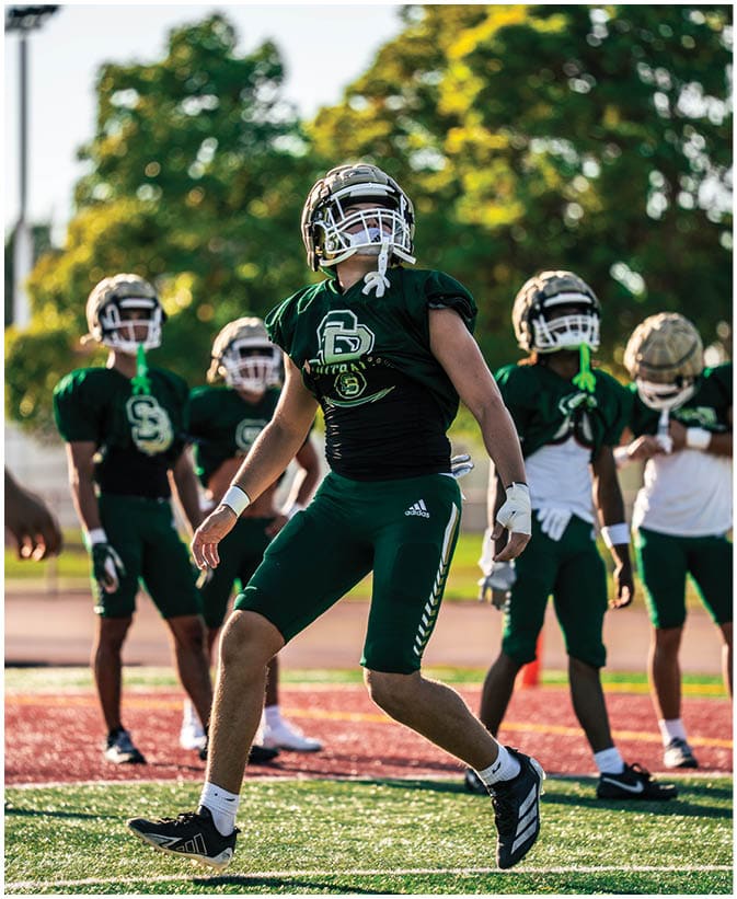 A person wearing a dark green football jersey and helmet looks upward with trees and a clear sky in the background. Description generated by AI