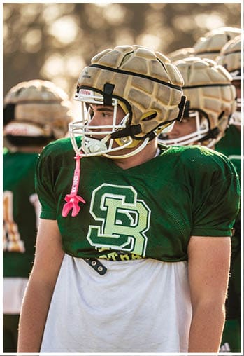 Two people wearing football helmets are shown with one person in clear focus and the other blurred in the background against a soft, natural backdrop. Description generated by AI