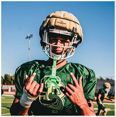 A person wearing a football helmet and green jersey stands outdoors under a clear blue sky with a light pole in the background. Description generated by AI