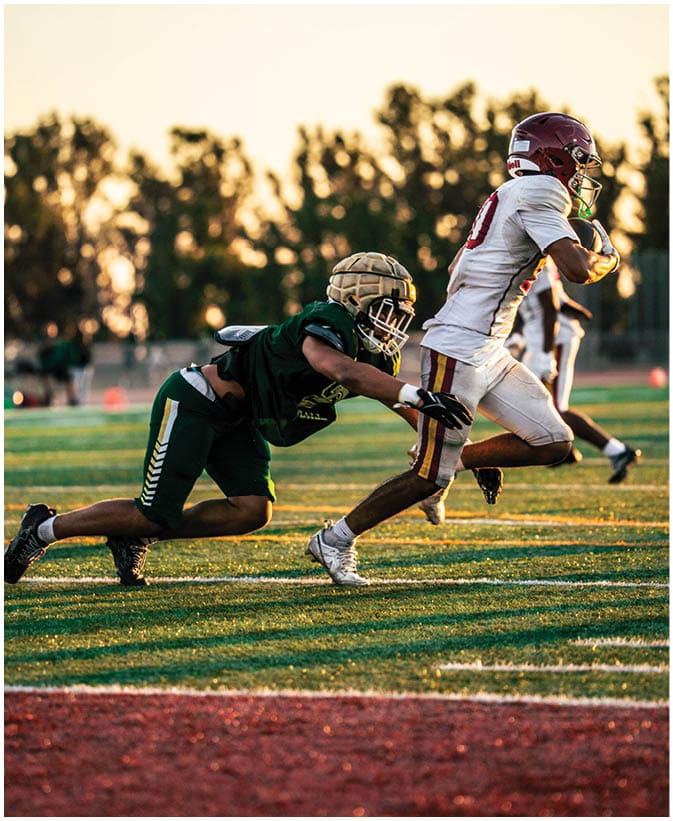 A person in a white football uniform is running with the ball while another person in a green uniform is attempting to tackle them on a football field during sunset. Description generated by AI