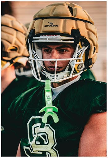 A person wearing a green football jersey and a tan padded helmet is standing with another similarly dressed person blurred in the background. Description generated by AI