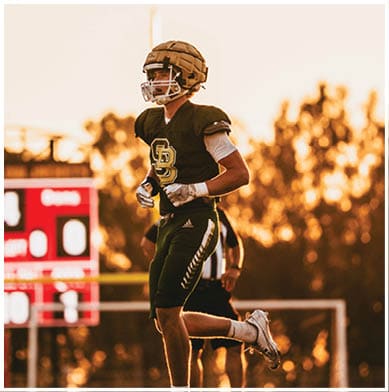 An adult wearing a football uniform and helmet is running on a field with a scoreboard and trees in the background during sunset. Description generated by AI