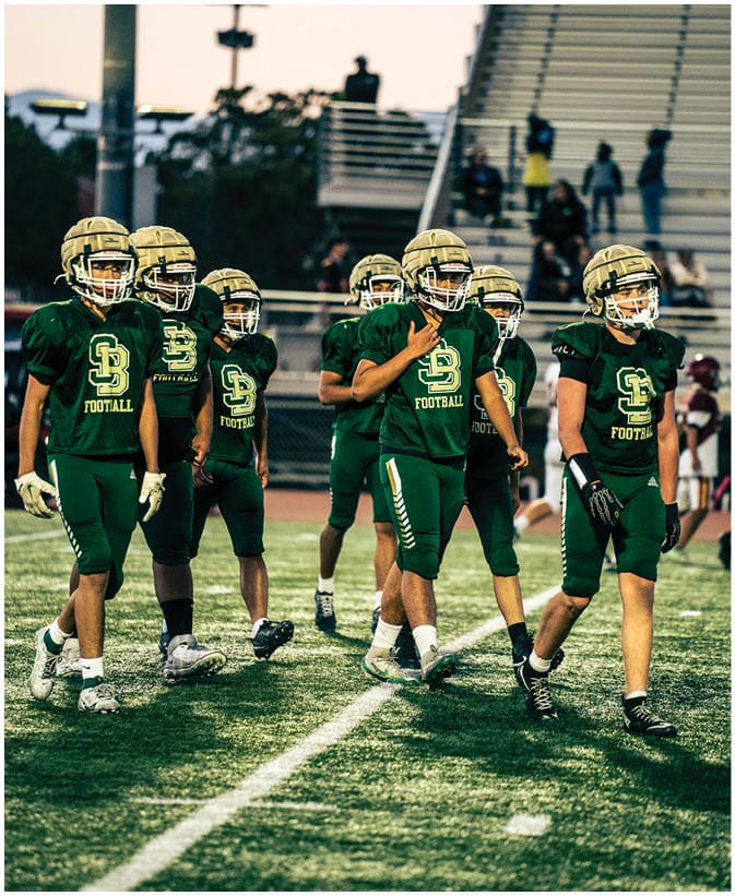 A group of people wearing green football uniforms and gold helmets are walking on a football field with bleachers and spectators in the background. Description generated by AI