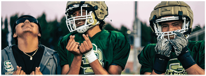 Three people wearing green football jerseys are standing together outdoors with two of them making hand gestures and one looking upward. Description generated by AI