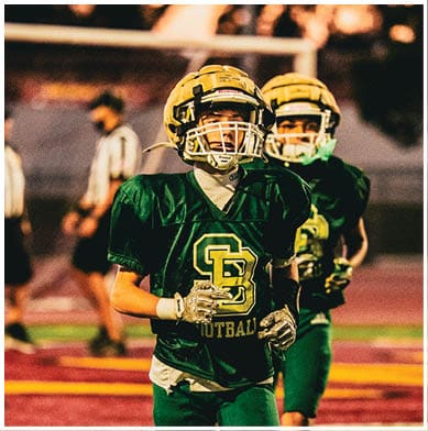 Two people wearing yellow helmets are seen from behind with a blurred outdoor background featuring a goalpost and trees. Description generated by AI